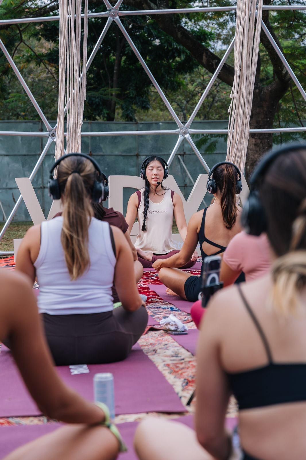 Letícia conduz uma meditação com fones de ouvido para um grupo sentado em tapetinhos, ao ar livre