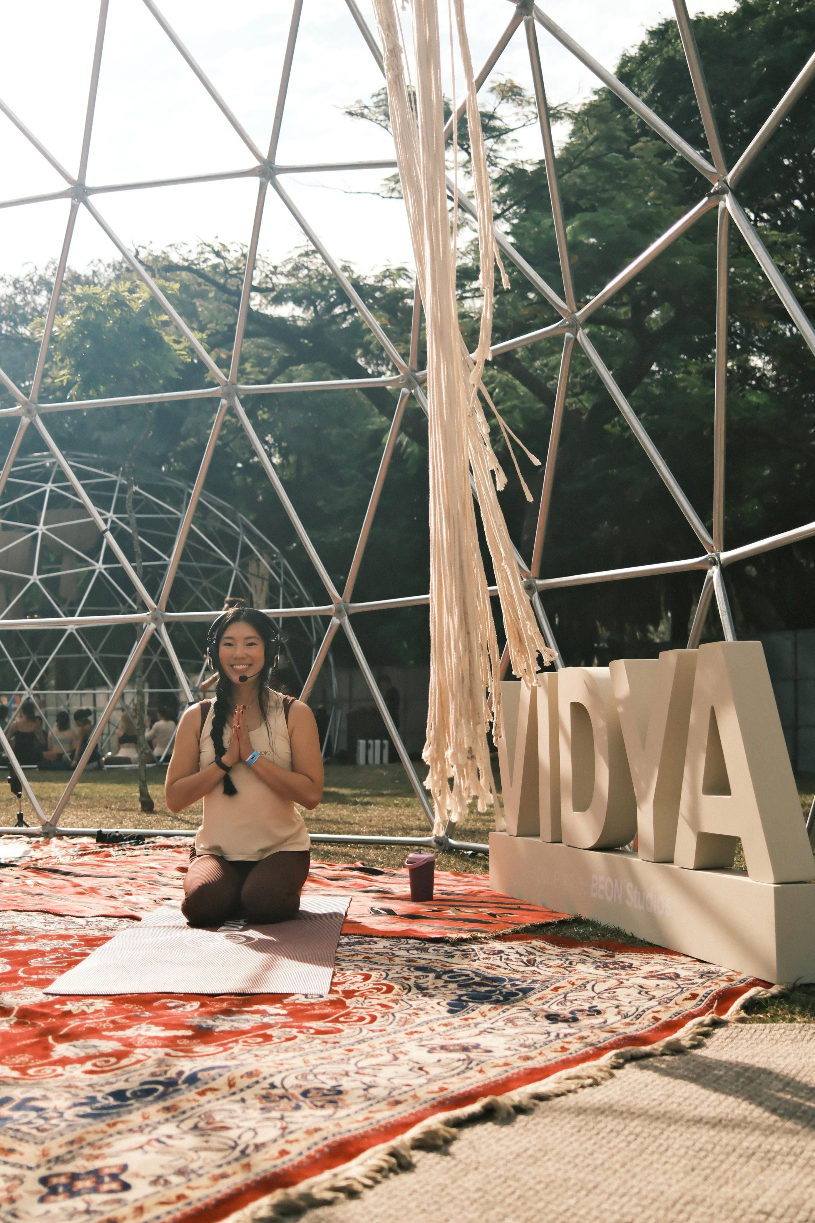 Letícia Yamane sorrindo, de mãos unidas, ajoelhada sobre tapetes persas sob um domo geodésico, no NOW Festival